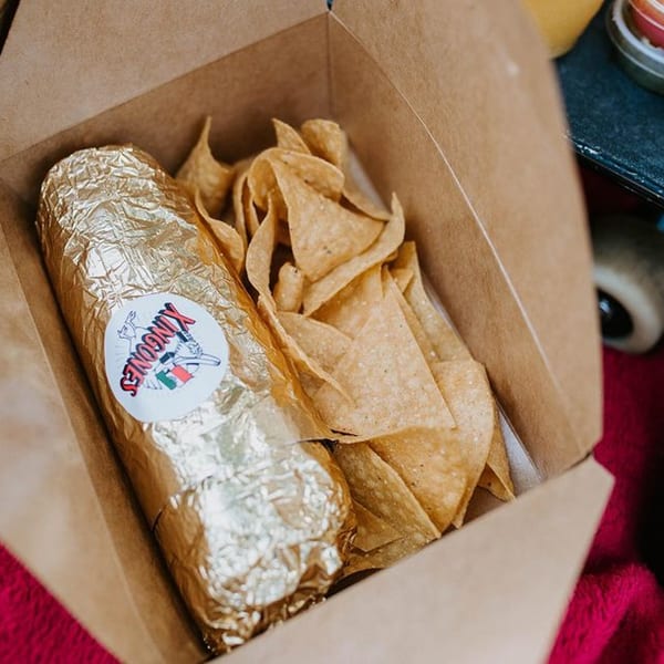 Xingones Cantina burrito wrapped in gold foil with a logo sticker, alongside tortilla chips in a takeout box.