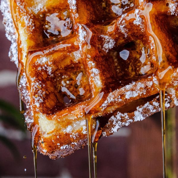 Close-up of a golden waffle, dusted with powdered sugar, with maple syrup dripping down.
