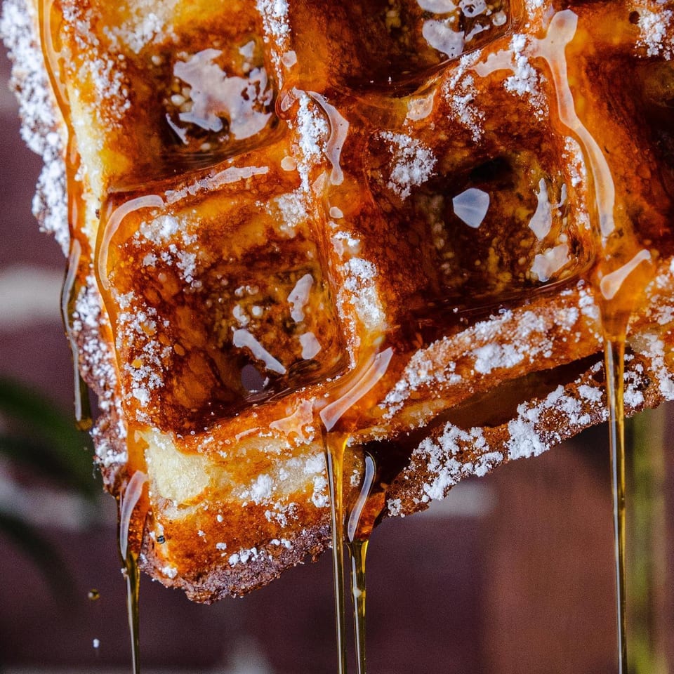 Close-up of a golden waffle, dusted with powdered sugar, with maple syrup dripping down.