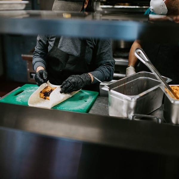 A chef wearing black gloves prepares a burrito on a green cutting board at Xingones Cantina.