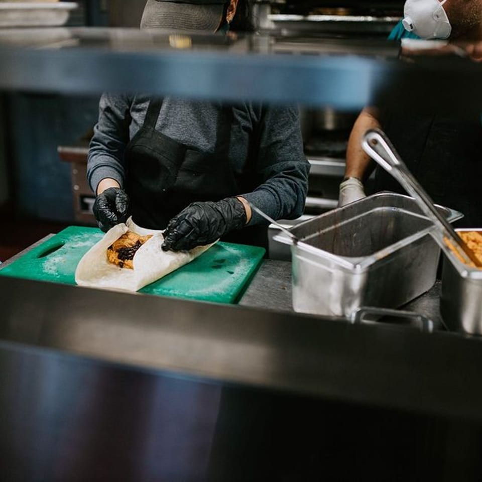 A chef wearing black gloves prepares a burrito on a green cutting board at Xingones Cantina.