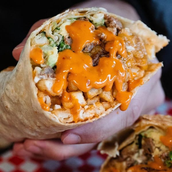 A hand holds a large, cut burrito from Xingones Cantina, filled with meat, fries, and orange sauce.