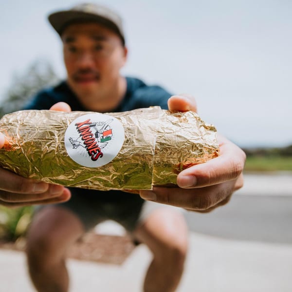 A person holds out a large burrito wrapped in gold foil with a "Xingones" sticker.