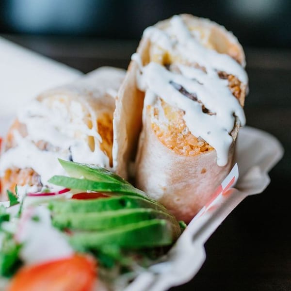 Two burritos drizzled with white sauce, served with sliced avocado and radish.