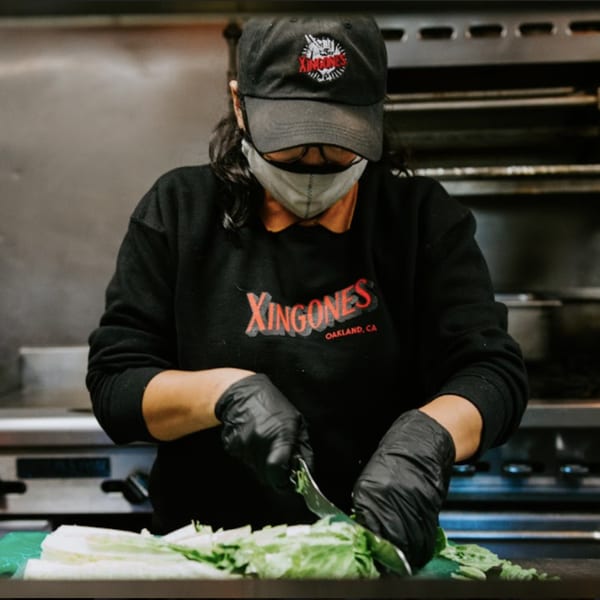A Xingones Cantina chef in a mask and gloves chops lettuce on a cutting board.