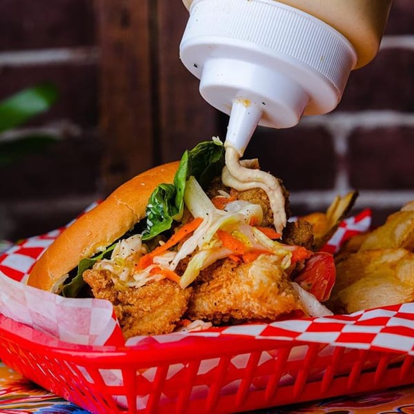 Sauce being squeezed onto a fried fish sandwich with slaw and chips in a red basket.
