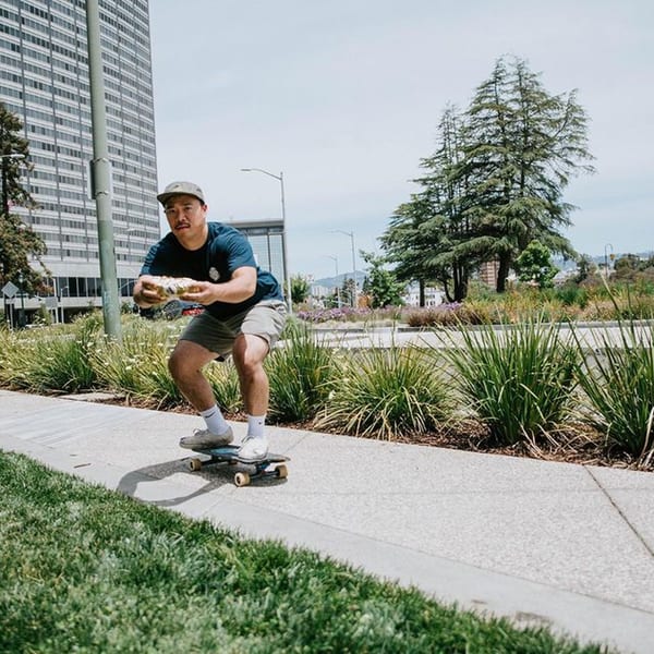 Man skateboarding on a sidewalk, holding a burrito, with a city building and trees in the background.