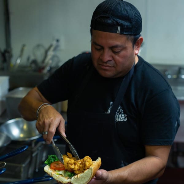 Chef in a black cap and apron assembling a Mexican sandwich with tongs in a kitchen.